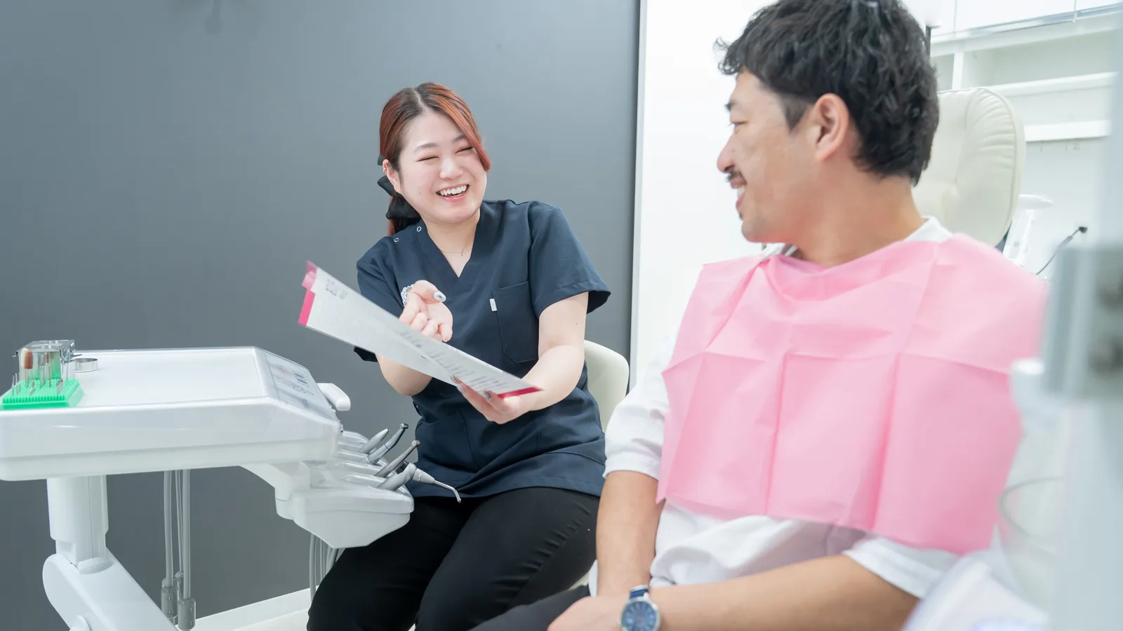 歯科医院で診療着の女性スタッフが笑顔で説明資料を見せながら、ピンクのエプロンを着けた男性患者と会話しているカウンセリング風景